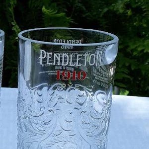 May include: Clear glass tumbler with embossed floral design. The glass features the "PENDLETON 1910" logo in white and red. The glass is on a light blue surface with green foliage in the background.