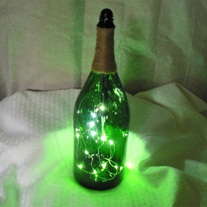 Puede incluir: Una botella de vidrio verde con una cuerda marrón envuelta alrededor del cuello. La botella está llena de luces blancas.
