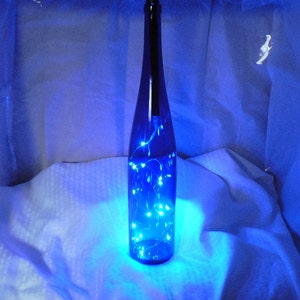 May include: A blue glass bottle with a string of blue fairy lights inside. The bottle is sitting on a white surface.