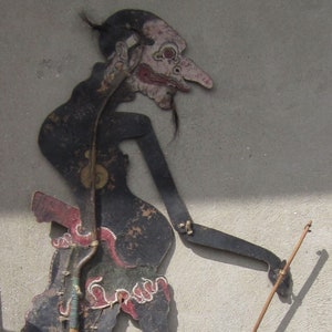 May include: A black and red painted wooden shadow puppet of a mythical creature with a long nose and beard. The puppet is attached to a wooden stick.