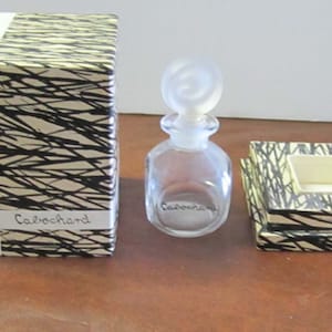 May include: A vintage perfume bottle with a clear glass body and a frosted glass stopper. The bottle is labeled "Cabochard" and is sitting on a white and black patterned box. The box is labeled "GRES PARIS".
