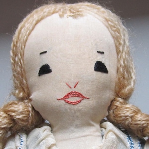 May include: A handmade cloth doll with a light beige face and blonde yarn hair styled in braids. The doll's facial features include embroidered black eyes, eyebrows, and a red stitched mouth. The doll is wearing a white dress.