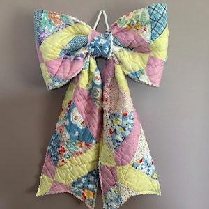 May include: A large decorative bow constructed from quilted fabric featuring a patchwork design. The textile displays a variety of floral and geometric patterns in pastel shades of pink, yellow, and blue, finished with a white scalloped edge trim.