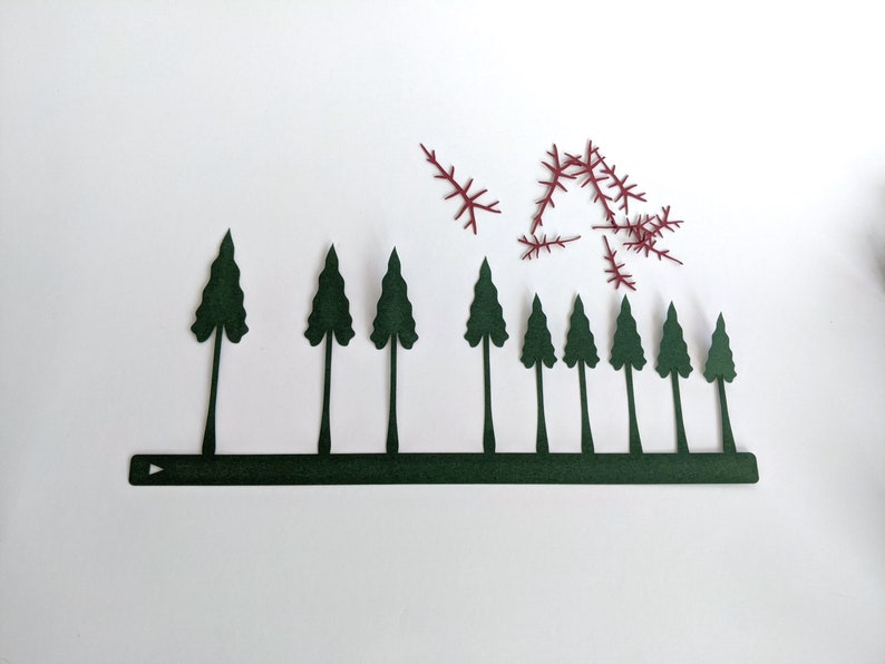May include: A row of ten dark green paper trees with a dark green paper base. There are several small, red paper sprigs scattered above the trees.