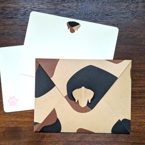 May include: Two white envelopes with a brown envelope with a camouflage pattern. The white envelopes have a paw print and a dog silhouette on them. The brown envelope has a dog silhouette on it.