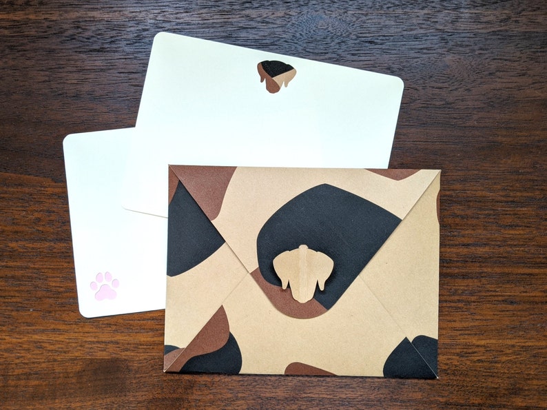 May include: Two white envelopes with a brown envelope with a camouflage pattern. The white envelopes have a paw print and a dog silhouette on them. The brown envelope has a dog silhouette on it.