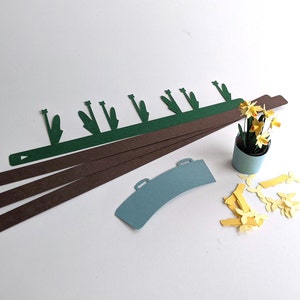 May include: A collection of paper cutouts for a craft project. Includes brown and green strips, a blue watering can shape, and a small pot with yellow paper flowers. The pieces are arranged on a white surface.