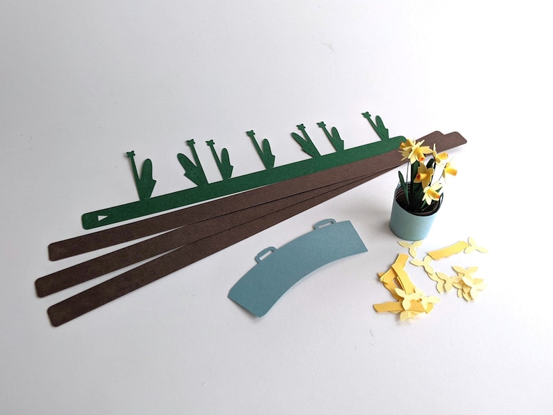 May include: A collection of paper cutouts for a craft project. Includes brown and green strips, a blue watering can shape, and a small pot with yellow paper flowers. The pieces are arranged on a white surface.