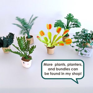 May include: A collection of paper plants, including a Venus flytrap, palm, and other greenery, in small pots. The plants are various shades of green and are arranged on a white surface. Text in a speech bubble reads: "More plants, planters, and bundles can be found in my shop!"