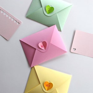 Op de afbeelding: Een verzameling kleurrijke papieren enveloppen en kaarten. De enveloppen zijn in roze, groen en geel, elk met een hartvormige sluiting. Sommige kaarten hebben hartvormige uitsparingen. E&eacute;n roze envelop heeft "Hi" op de hartsluiting, een andere "Hey".