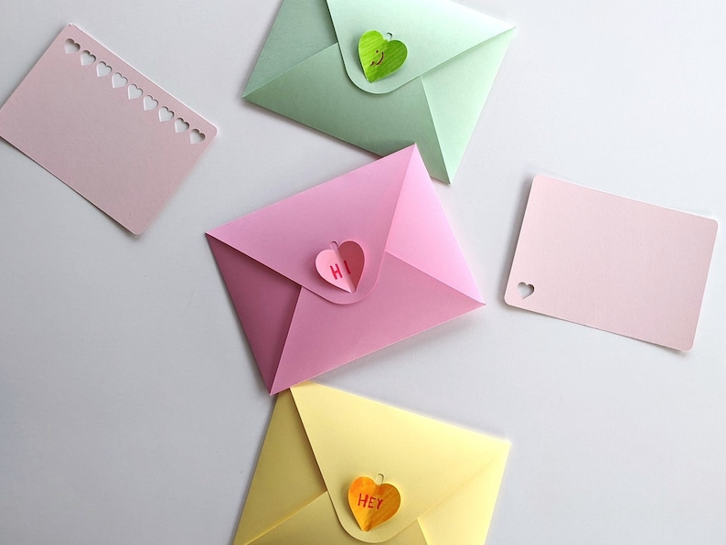 Op de afbeelding: Een verzameling kleurrijke papieren enveloppen en kaarten. De enveloppen zijn in roze, groen en geel, elk met een hartvormige sluiting. Sommige kaarten hebben hartvormige uitsparingen. E&eacute;n roze envelop heeft "Hi" op de hartsluiting, een andere "Hey".