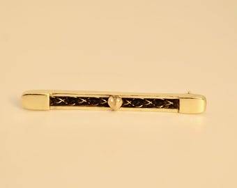 May include: A gold-toned bar pin with a central woven design and a small, heart-shaped accent. The pin has a rectangular shape with rounded ends. The woven section is dark, contrasting with the gold color.