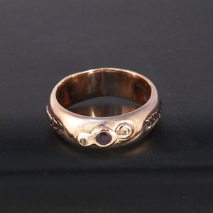 May include: A rose gold ring with a rounded band. The ring features a central dark purple gemstone, flanked by two smaller clear stones and decorative black accents. The ring is set against a dark background.