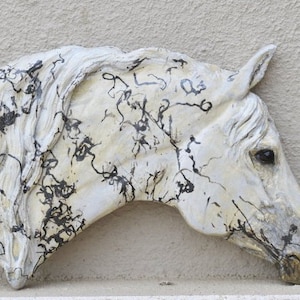 May include: A white horse head sculpture with black abstract markings. The sculpture is mounted on a light beige wall. The horse's mane and face are detailed, with a slightly weathered appearance. The sculpture is a decorative piece.