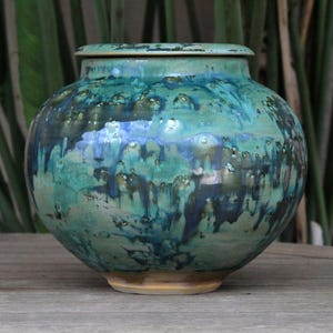 May include: A glazed ceramic jar with a lid, in a rounded shape. The jar is predominantly teal and blue, with brown accents and a glossy finish. It sits on a light brown base. The background is blurred, with green foliage visible.