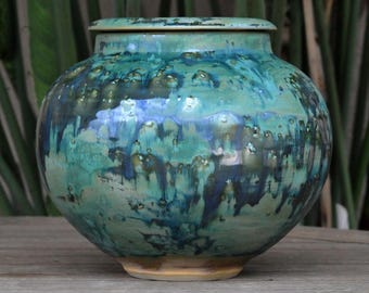 Op de afbeelding: Een keramische pot met deksel, met een afgeronde vorm en een getextureerd oppervlak. De pot is geglazuurd in tinten van turquoise, blauw en bruin, met een glanzende afwerking. De pot staat op een lichtbruine basis.