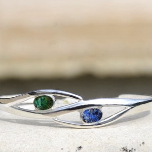 May include: Silver cuff bracelet with a wave design and two oval-shaped gemstones. One gemstone is green and the other is blue.