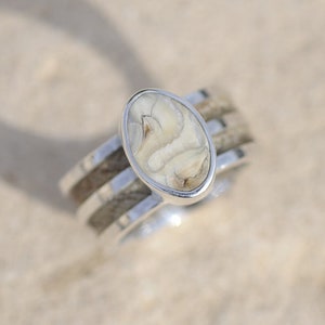 Op de afbeelding: Een zilveren ring met een witte ovale steen in het midden. De ring heeft een drievoudig bandontwerp met een houtstructuur.