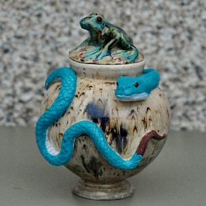 Könnte beinhalten: Eine Keramikvase mit Deckel, auf dem eine Froschfigur sitzt. Eine türkisfarbene Schlange windet sich um die Vase, mit einem Schlangenkopf an der Seite. Die Vase hat ein gesprenkeltes, erdiges Farbschema mit Braun- und Cremetönen.