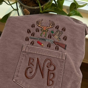 Puede incluir: Una camiseta de color malva con bolsillo y un bordado que muestra una cornamenta de ciervo con un lazo verde, un pato real en vuelo, una escopeta, un cartucho rojo y huellas de pezuñas marrones. El bolsillo presenta una letra N bordada.