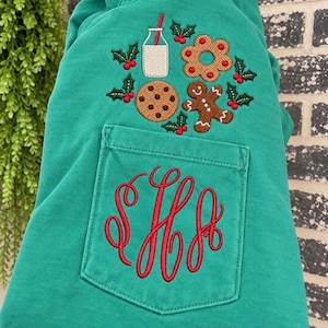 May include: A green sweatshirt featuring an embroidered design of a glass of milk, a cookie, a gingerbread man, and holly leaves above a chest pocket. The pocket displays a large, red, cursive monogram with three intertwined letters.