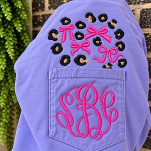May include: A light purple sweatshirt featuring a chest pocket with an embroidered hot pink monogram of the letters SBC. Above the pocket is an embroidered pattern of black and tan leopard print spots interspersed with four hot pink bows.