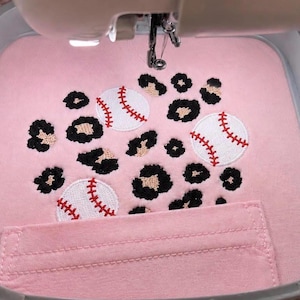 May include: A light pink fabric piece secured in an embroidery hoop, featuring an embroidered pattern of three white baseballs with red stitching interspersed among black and tan leopard print spots, positioned above a horizontal fabric pocket.