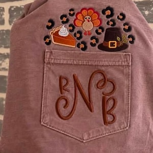 May include: A mauve-coloured shirt featuring an embroidered pocket with the monogram letters R, N, and B. Above the pocket, an embroidered design displays a slice of pumpkin pie, a turkey, and a pilgrim hat, all surrounded by leopard print spots.