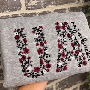 May include: A heather grey sweatshirt featuring embroidered floral patterns in shades of dark red, white, and black. The embroidery forms the letters U and A, with the word razorbacks stitched in a cursive black font along the right side.