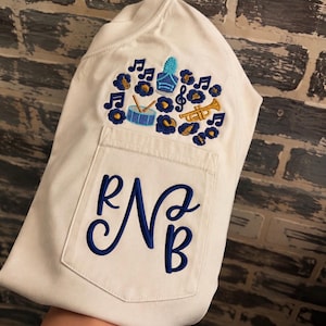 May include: A cream-colored t-shirt featuring a chest pocket embroidered with the dark blue monogram letters R, N, and B. Above the pocket, a colorful embroidered design displays musical instruments including a drum and trumpet with notes.