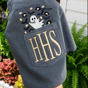May include: A charcoal gray t-shirt featuring a chest pocket embroidered with the letters HHS in tan thread. Above the pocket is a graphic design of a white ghost surrounded by black bats and tan leopard print spots. The text DO NOT COPY is visible.