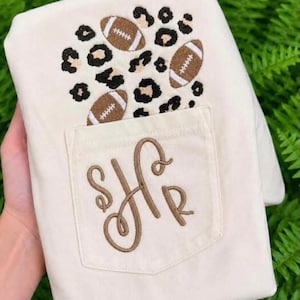 Puede incluir: Una camiseta color crema con un diseño bordado de manchas de leopardo marrones y tres balones de fútbol americano marrones sobre un bolsillo en el pecho. El bolsillo muestra un monograma grande bordado en cursiva marrón con las letras S, H y R.