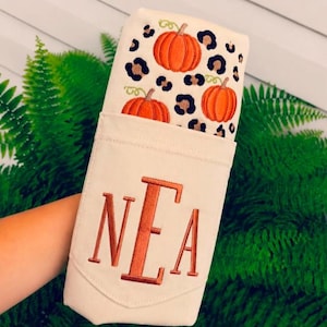 May include: A cream-colored fabric pouch featuring an embroidered pattern of orange pumpkins and black and tan leopard spots. The front of the pouch displays the embroidered monogram letters NEA in a bold, dark orange font with a serif typeface.