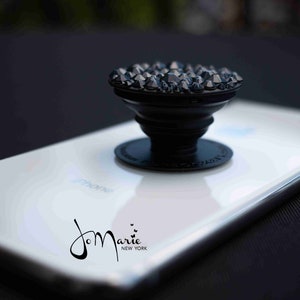Peut inclure: Une poignée de téléphone noire avec un design en cristal noir scintillant. La poignée est fixée à un smartphone blanc avec le texte "J.Marie New York" imprimé au dos.