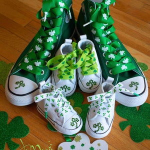 May include: A set of four green and white trainers with shamrock designs. The two larger trainers have green laces with shamrock ribbon, while the two smaller trainers have green and white shamrock ribbon laces. The trainers are on a wooden surface with green shamrock cutouts.