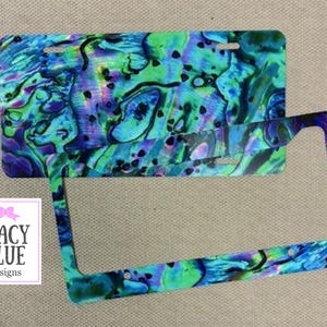 May include: License plate and frame with a vibrant, swirling pattern in shades of blue, green, and purple. The design resembles abalone shell. The Macy Blue Designs logo is in the lower left corner.