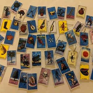 Puede incluir: Una colección de aretes con forma de cartas de Lotería coloridas. Las cartas presentan varias imágenes, incluyendo una pera, un camarón, una palmera, una rana, una calavera, una campana, una sandía y una mano. Los aretes están hechos de metal y tienen un acabado plateado.