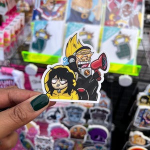 May include: A sticker featuring three anime characters. The character on the left is wearing a yellow raincoat and has a surprised expression. The character in the middle is wearing a black and white outfit and has a microphone. The character on the right is wearing a black and white outfit and has a surprised expression.