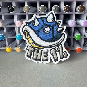 May include: Embroidered patch featuring a blue and white turtle shell design with the text "THE 1%". The patch has a white border and is set against a background of art markers.