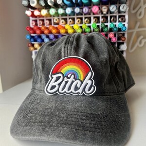 May include: A grey, distressed baseball cap with a curved peak. The cap has a rainbow patch with the word "Bitch" in white script. The background shows a white organiser filled with colourful markers.