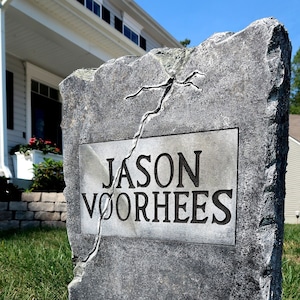 May include: A weathered, gray tombstone with the name "JASON VOORHEES" inscribed on a rectangular plaque. A large crack runs across the stone. The tombstone is set in green grass, with a house visible in the background.