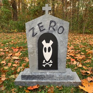 以下が含まれることがあります： 「ZERO」の文字が書かれた、黒いアーチの上に灰色の石のような墓石。アーチの中には、白い幽霊のような姿と交差した骨があります。墓石の上部には十字架があり、長方形の台座の上に立っています。