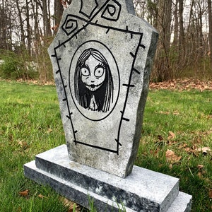 May include: A grey stone tombstone with a black and white drawing of a girl with long hair and large eyes. The tombstone has a stitched border and is decorated with a few simple designs.