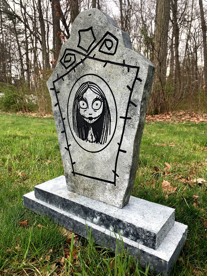 May include: A grey stone tombstone with a black and white drawing of a girl with long hair and large eyes. The tombstone has a stitched border and is decorated with a few simple designs.