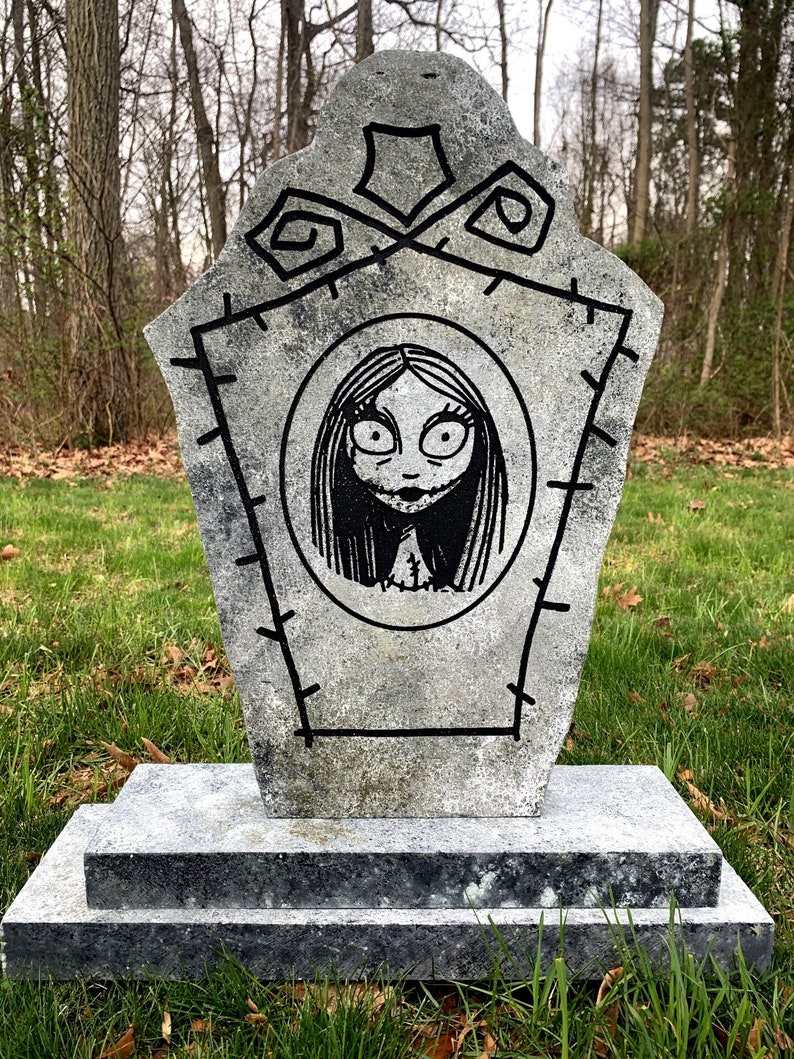 May include: A grey stone tombstone with a black and white drawing of a girl with large eyes. The tombstone has a black border and a stitched design. The tombstone is on a two-tiered base.