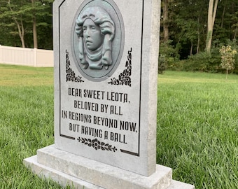 May include: A grey stone tombstone with a carved portrait of a woman with curly hair. The inscription reads "DEAR SWEET LEOTA, BELOVED BY ALL IN REGIONS BEYOND NOW, BUT HAVING A BALL".
