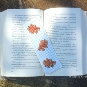 Könnte beinhalten: Nahaufnahme eines Buches mit einem Lesezeichen, das eine handgezeichnete Illustration von Herbstblättern zeigt. Das Lesezeichen besteht aus kariertem Papier mit weißem Hintergrund. Das Buch ist auf einer Seite mit polnischem Text aufgeschlagen.