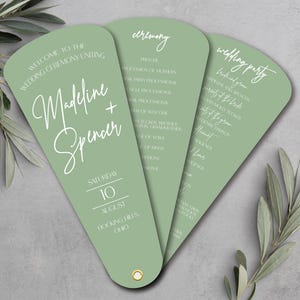 May include: Sage green wedding program fans with white calligraphy text. The fans display the names "Madeline + Spencer", ceremony details, and wedding party information. The date is August 10th, in Hocking Hills, Ohio.