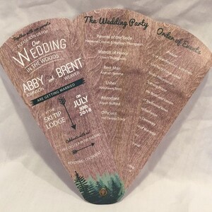 May include: A wedding invitation in the form of a wooden fan with a rustic design. The invitation features the couple's names, wedding date, and location. The back of the fan lists the wedding party and order of events.
