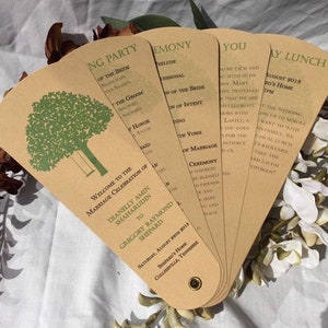 May include: A set of brown paper fans with a green tree and swing design. The fans have text on them that reads "Welcome to the Marriage Celebration of Dianelly Amin Shaharudin to Gregory Raymond Shepard" and includes the date and location of the wedding. The fans are laid out on a white surface with white flowers.